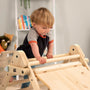 A young child climbs on the TP Active-Tots Pikler Style Wooden Climbing Triangle by TP Toys, an FSC® certified Montessori toy, with books and toys visible in the background.