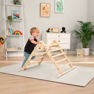 A toddler stands and climbs on the TP Toys TP Active-Tots Pikler Style Wooden Climbing Triangle - FSC® certified in a playroom featuring a bookshelf, dresser, toys, and wall art in the background.