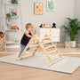 A toddler stands and climbs on the TP Toys TP Active-Tots Pikler Style Wooden Climbing Triangle - FSC® certified in a playroom featuring a bookshelf, dresser, toys, and wall art in the background.