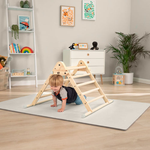 A toddler crawls under the TP Toys Active-Tots Pikler Style Wooden Climbing Triangle (FSC® certified) on a play mat in a bright, modern playroom with shelves, plants, and children's artwork.