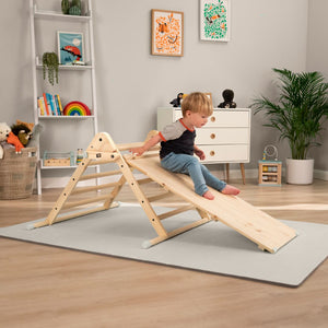 A young child sits atop the TP Toys TP Active-Tots Pikler Style Wooden Climbing Triangle & Slide (FSC® certified) in a playroom with toys, a plant, and framed art on the wall.