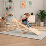 A young child sits atop the TP Toys TP Active-Tots Pikler Style Wooden Climbing Triangle & Slide (FSC® certified) in a playroom with toys, a plant, and framed art on the wall.