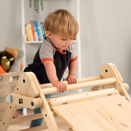 A young child climbs on the TP Toys TP Active-Tots Pikler Style Wooden Climbing Triangle & Slide (FSC® certified) in a room with shelves and books in the background.