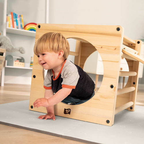 A young child crawls through the TP Toys TP Active-Tots Pikler Style Wooden Climbing Cube—FSC® certified—on a padded floor in a bright, child-friendly room.
