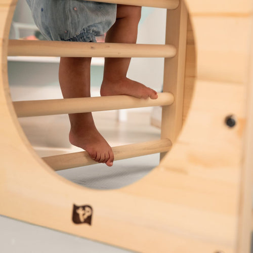 A child climbs barefoot on the TP Toys Active-Tots Pikler Style Wooden Climbing Cube (FSC® certified), viewed through its circular opening, with horizontal bars forming part of the wooden play structure.