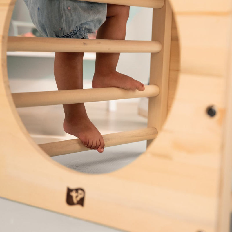 A child climbs barefoot on the TP Toys Active-Tots Pikler Style Wooden Climbing Cube (FSC® certified), viewed through its circular opening, with horizontal bars forming part of the wooden play structure.