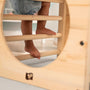A child climbs barefoot on the TP Toys Active-Tots Pikler Style Wooden Climbing Cube (FSC® certified), viewed through its circular opening, with horizontal bars forming part of the wooden play structure.