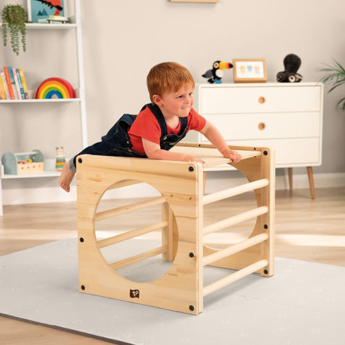 A young child in a red shirt and overalls climbs on the TP Active-Tots Pikler Style Wooden Climbing Cube by TP Toys in a brightly lit, neatly organized playroom.