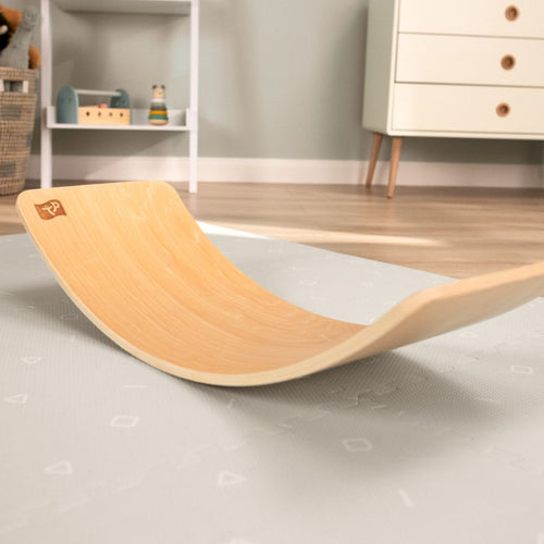 The TP Toys Active-Tots Pikler Style Wooden Balance Board - FSC® certified rests on a light gray play mat in a children’s room, promoting gross motor skills, with shelves and a dresser in the background.