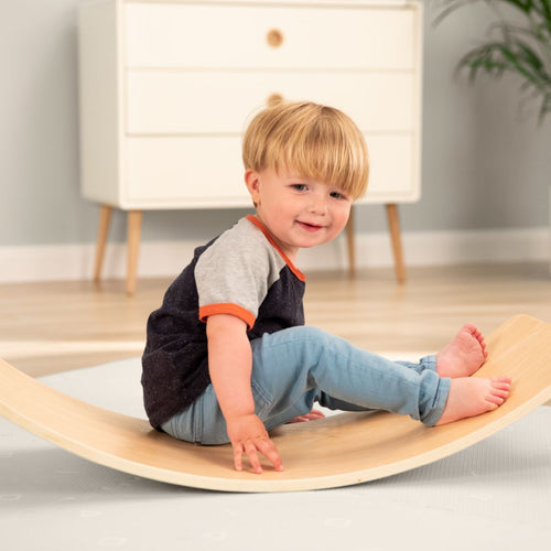 A young child balances on the TP Toys TP Active-Tots Pikler Style Wooden Balance Board (FSC® certified) in a modern, light-filled room with a dresser, building gross motor skills through play.