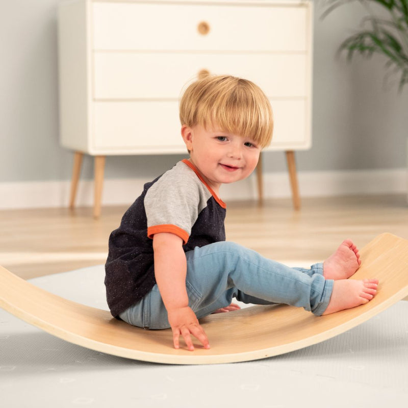 A young child balances on the TP Toys TP Active-Tots Pikler Style Wooden Balance Board (FSC® certified) in a modern, light-filled room with a dresser, building gross motor skills through play.