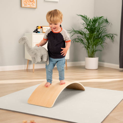 A young child stands barefoot on the TP Active-Tots Pikler Style Wooden Balance Board by TP Toys, holding a stuffed dinosaur in a bright, tidy room—a fun Montessori-inspired way to build gross motor skills with this FSC® certified toy.
