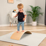 A young child stands barefoot on the TP Active-Tots Pikler Style Wooden Balance Board by TP Toys, holding a stuffed dinosaur in a bright, tidy room—a fun Montessori-inspired way to build gross motor skills with this FSC® certified toy.