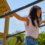 A young girl balances on the TP Toys TP Skywood Wooden Clatter Bridge—FSC® certified—at a playground, wearing a white sleeveless top and denim shorts, surrounded by trees and blue sky.
