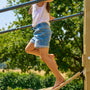A child climbs the TP Toys Skywood Wooden Clatter Bridge—FSC® certified—on an outdoor playground, gripping metal bars and wearing a white shirt with denim shorts.
