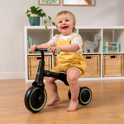 A smiling toddler in yellow overalls sits on a TP Toys Zoomee Balance Bike to Trike indoors, supporting child development, with shelves and baskets in the background.