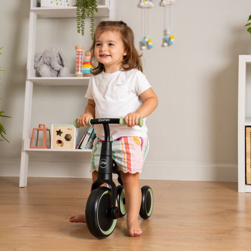 A young child indoors smiles while holding the handlebars of a Zoomee Balance Bike to Trike by TP Toys—a fun way to support early development. Toy- and decoration-filled shelves are visible in the background.