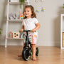 A young child indoors smiles while holding the handlebars of a Zoomee Balance Bike to Trike by TP Toys—a fun way to support early development. Toy- and decoration-filled shelves are visible in the background.