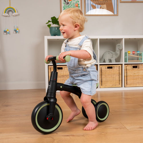 A toddler in denim overalls rides the TP Toys Zoomee Balance Bike to Trike indoors on a wooden floor, building confidence and supporting child development.