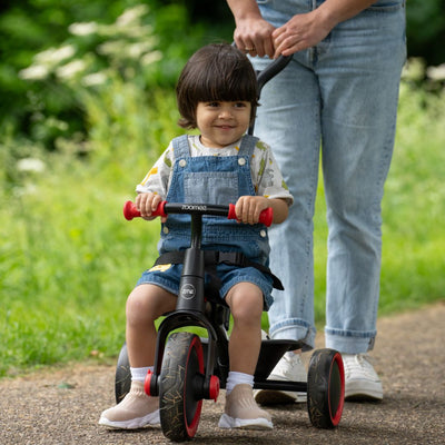 A toddler in denim overalls rides the TP Toys Zoomee 2 in 1 Trike – Firecracker, featuring red handles, while an adult pushes from behind on a paved outdoor path.