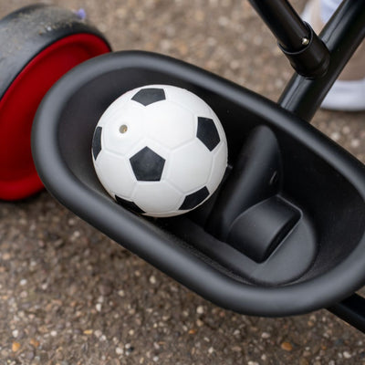 A small black and white soccer ball rests in the storage basket of a TP Toys Zoomee 2 in 1 Trike – Firecracker with an adjustable handle, positioned on a paved surface.