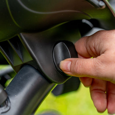 A hand turns the black dial on the adjustable handle of the TP Toys Zoomee 2 in 1 Trike – Firecracker, with a green blurred background highlighting its outdoor use.