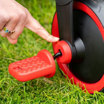 A hand pushes the Zoomee 2 in 1 Trike – Firecracker by TP Toys, featuring a red pedal and black-and-red wheel on grass—an exciting trike made for kids’ outdoor adventures.