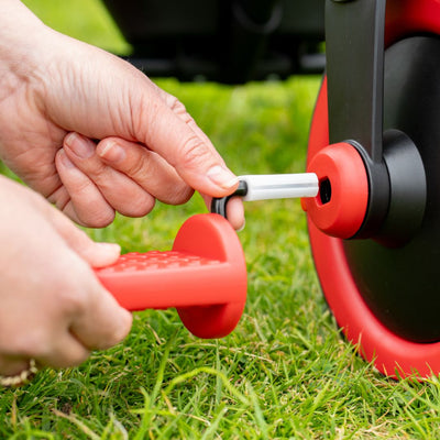 Someone’s hands assemble a Zoomee 2 in 1 Trike – Firecracker by TP Toys, attaching a red and black wheel to an axle outdoors on the grass, preparing the trike for play.