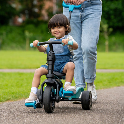 A young child rides the TP Toys Zoomee 4 in 1 Trike - Dino Discovery on a path as an adult in jeans and sneakers pushes from behind in a grassy park.