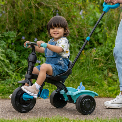 A young child in denim overalls rides the TP Toys Zoomee 4 in 1 Trike - Dino Discovery, being guided outdoors on a paved path by an adult using the push handle.
