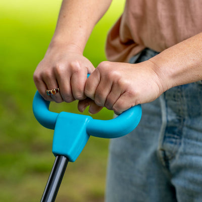 A person in jeans holds the handle of a Zoomee 4 in 1 Trike - Dino Discovery by TP Toys outdoors, with grass and a hint of dinosaur print on their shirt visible in the background.