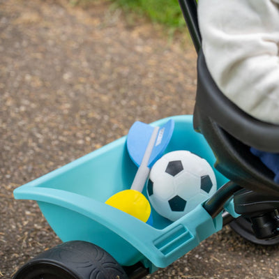 A close-up of the Zoomee 4 in 1 Trike - Dino Discovery by TP Toys, attached to a stroller, carrying a toy soccer ball, small bat, and yellow scoop on a paved path.
