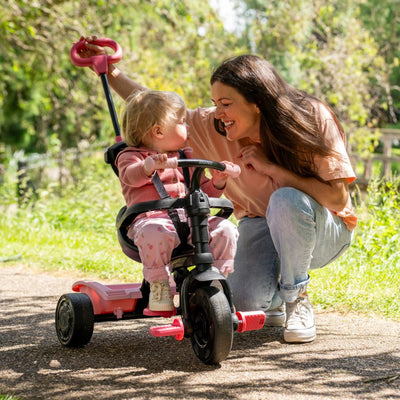 A woman squats next to a smiling toddler seated on the TP Toys Zoomee 4 in 1 Trike - Unicorn Dream with an adjustable UV sunshade, surrounded by lush greenery and sunlight.
