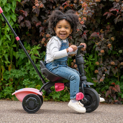 A young child smiles while sitting on the Zoomee 4 in 1 Trike - Unicorn Dream by TP Toys, featuring a push handle, outdoors in front of vibrant green and burgundy foliage.