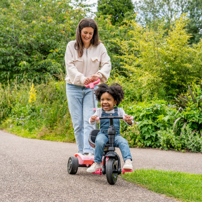 An adult pushes a smiling child riding the Zoomee 4 in 1 Trike - Unicorn Dream by TP Toys, featuring an adjustable UV sunshade, along a park path surrounded by lush greenery.