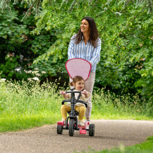 A woman pushes a smiling child on the TP Toys Zoomee 4 in 1 Trike – Unicorn Dream, featuring an adjustable UV sunshade, as they glide along a paved path in a green, leafy park.