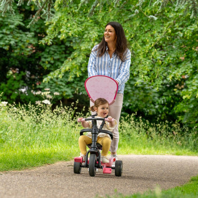 A woman pushes a smiling child on the TP Toys Zoomee 4 in 1 Trike – Unicorn Dream, featuring an adjustable UV sunshade, as they glide along a paved path in a green, leafy park.