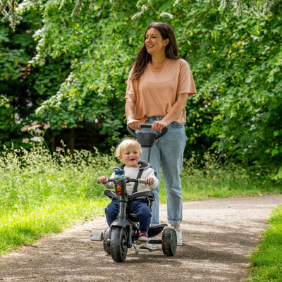 A woman pushes a smiling toddler on the Zoomee 4 in 1 Deluxe Trike - Grey Storm by TP Toys, featuring an adjustable seat, along a shaded path in a green park.