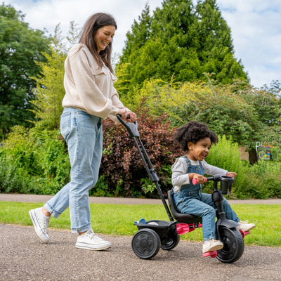 A woman and child share a joyful outdoor moment as she pushes the Zoomee 4 in 1 Deluxe Trike - Pink Dusk by TP Toys.