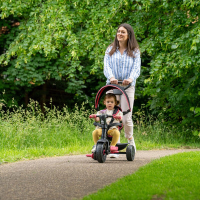 A woman pushes a young child on the Zoomee 4 in 1 Deluxe Trike by TP Toys in Pink Dusk along a paved path lined with green grass and trees.