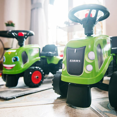 Two Falk Claas Baby Tractors by TP Toys, featuring green bodies, black wheels, and red accents, are indoors on a tiled floor with sunlight streaming in through a window in the background.