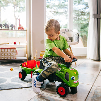 A young boy sits on a TP Toys Falk Claas Baby Tractor with a red trailer indoors, looking down to his left. Toys and furniture are visible in the background near large windows.