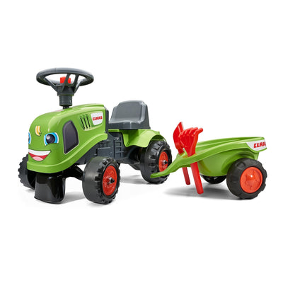 The Falk Claas Baby Tractor by TP Toys features a cheerful green design with a smiling face, plus a matching trailer carrying a red toy rake and shovel.
