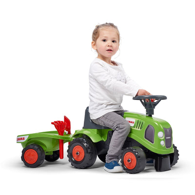 A young child sits on a TP Toys Falk Claas Baby Tractor with matching trailer, holding the steering wheel and facing slightly to the side.