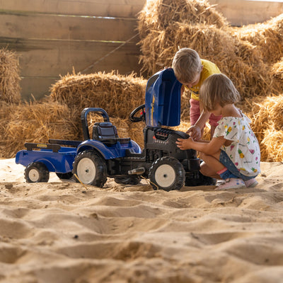 Two young children examine the engine of a Falk New Holland Tractor with Trailer and Opening Bonnet by TP Toys in a sandy barn area, with hay bales in the background.