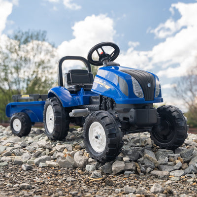 A Falk New Holland Tractor with Trailer and Opening Bonnet by TP Toys sits on gravel outdoors, with trees and a partly cloudy sky in the background.