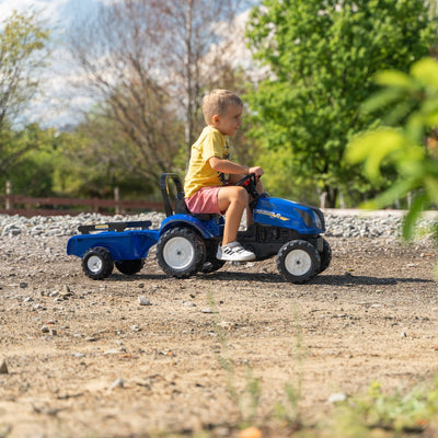 A young boy in a yellow shirt and pink shorts rides a TP Toys Falk New Holland Tractor with Trailer and Opening Bonnet on a gravel path outdoors.