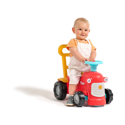 A toddler sits on the Falk Maurice Vintage Baby Red Tractor by TP Toys, a red and blue ride-on with large black wheels—ideal as a riding toy or walking aid to help little ones learn to walk—against a white background.