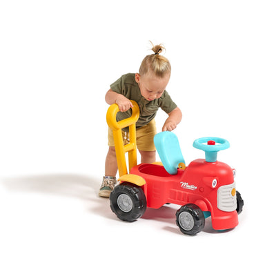 A young child stands beside the TP Toys Falk Maurice Vintage Baby Red Tractor, holding the yellow walking aid bar and lifting the blue seat lid.
