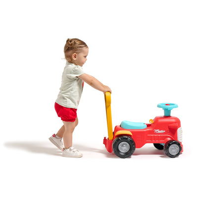 A young child uses the TP Toys Falk Maurice Vintage Baby Red Tractor as a walking aid, pushing the bright red ride-on with blue and yellow accents against a plain white background.